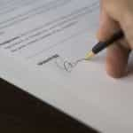 A person’s hand holding a pen and signing a written contract on paper with an official signature.