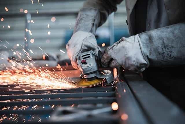 A worker using a blade saw on a piece of metal in a factory, with sparks flying out from the blade grinding against the metal.