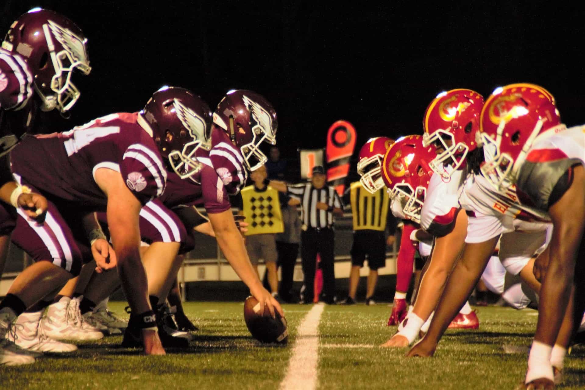 Two rows of American football players on opposing teams facing off one another at the beginning of a game on an outdoor field, with a referee off in the background standing in between the two.