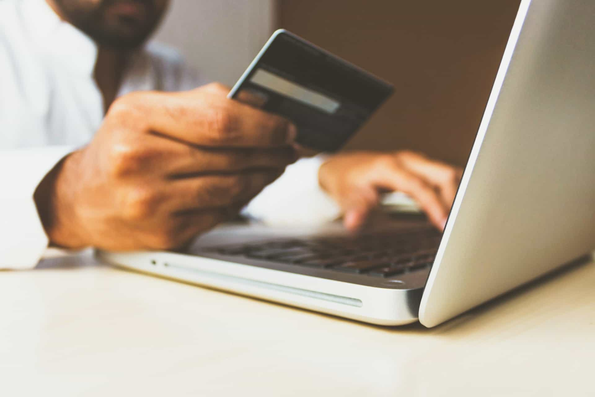 A person sitting at their laptop while holding a credit card in one hand, inputting information from the card onto a website or application on the laptop.