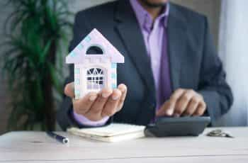 A man in a suit holding a small model house in the palm of his hand.