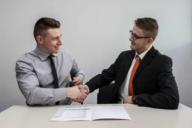 Two men in suits sitting down and facing one another while shaking each others’ hands.